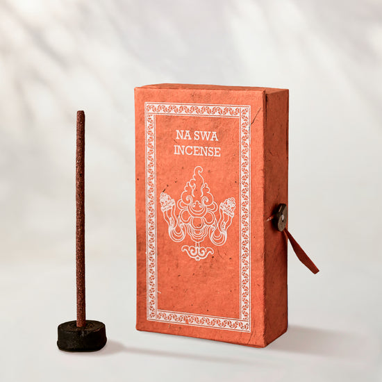 Na Swa Incense Sticks, box of 40: coral Stupa paper box with white print beside a stick standing in a small holder.
