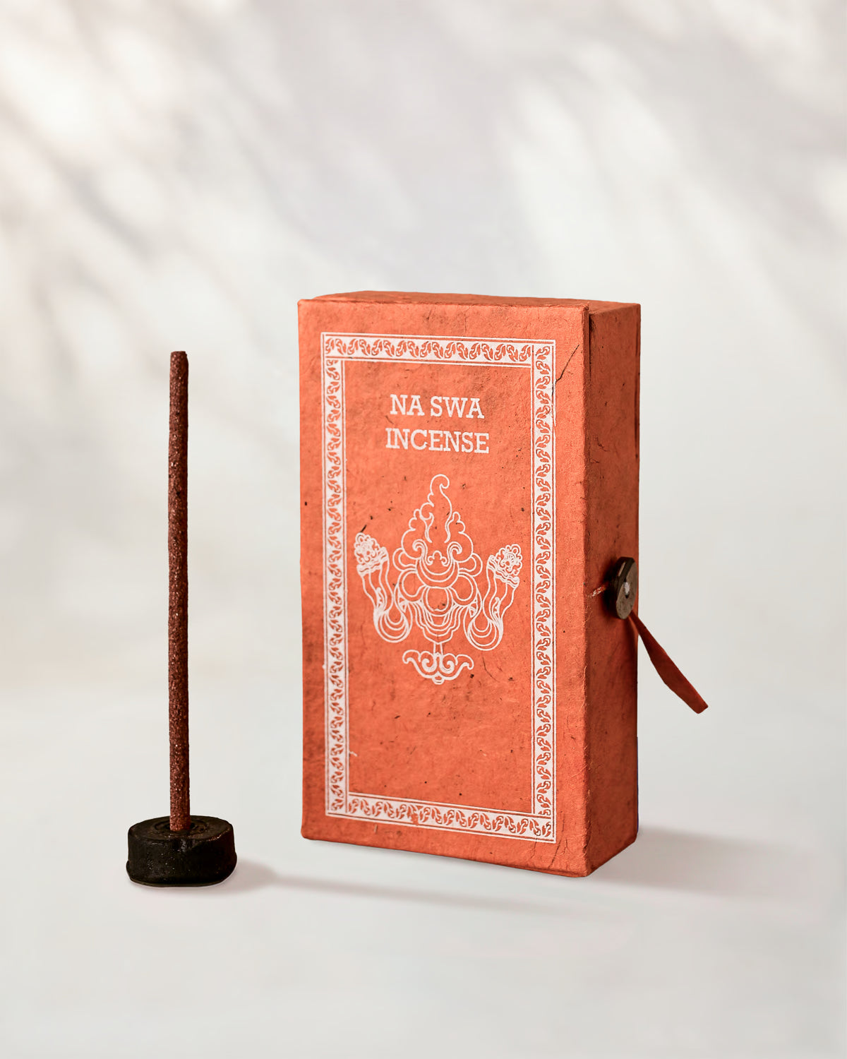 Na Swa Incense Sticks, box of 40: coral Stupa paper box with white print beside a stick standing in a small holder.