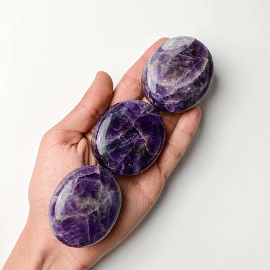 Three polished amethyst palm stones resting in an open hand against a white background.