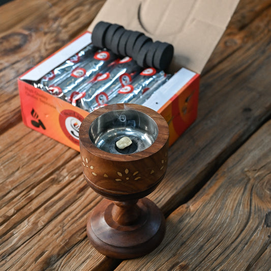 Wooden resin burner holding a charcoal tablet in front of an open box of charcoal rolls, on a wooden surface.