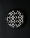 Round stone incense stick holder carved with a Flower of Life pattern, seen from above on a dark background.