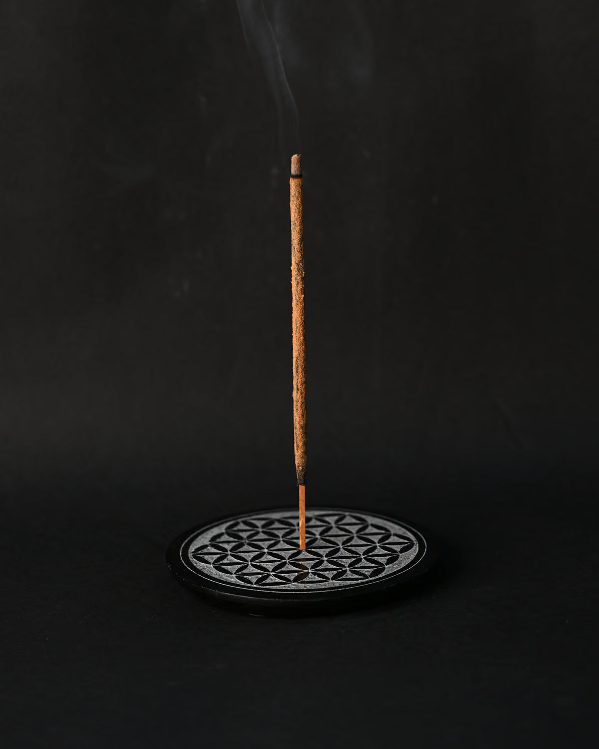 Incense stick burning upright in the round Flower of Life stone holder, smoke rising against a dark background.