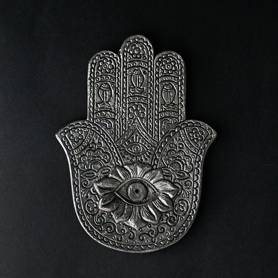 Hand-shaped incense stick holder in silver-toned metal with an eye motif at its centre, seen from above on a dark background.