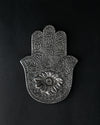 Hand-shaped incense stick holder in silver-toned metal with an eye motif at its centre, seen from above on a dark background.