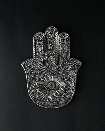 Hand-shaped incense stick holder in silver-toned metal with an eye motif at its centre, seen from above on a dark background.