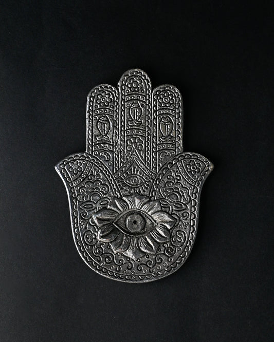 Hand-shaped incense stick holder in silver-toned metal with an eye motif at its centre, seen from above on a dark background.