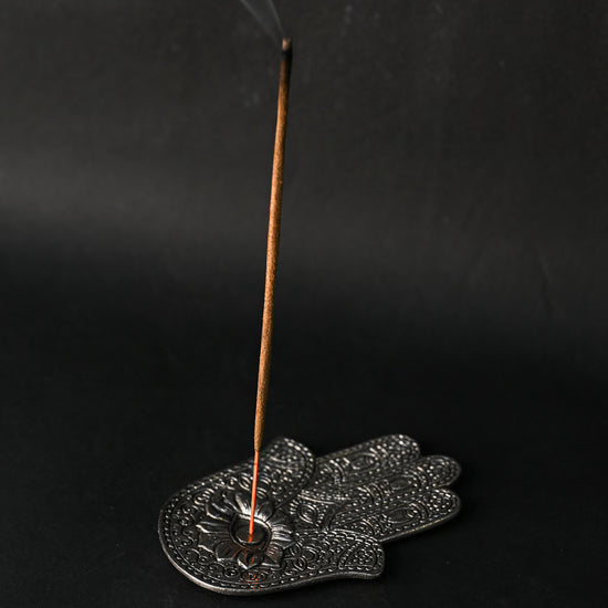 Incense stick burning upright in the hand-shaped metal incense holder, smoke rising against a dark background.