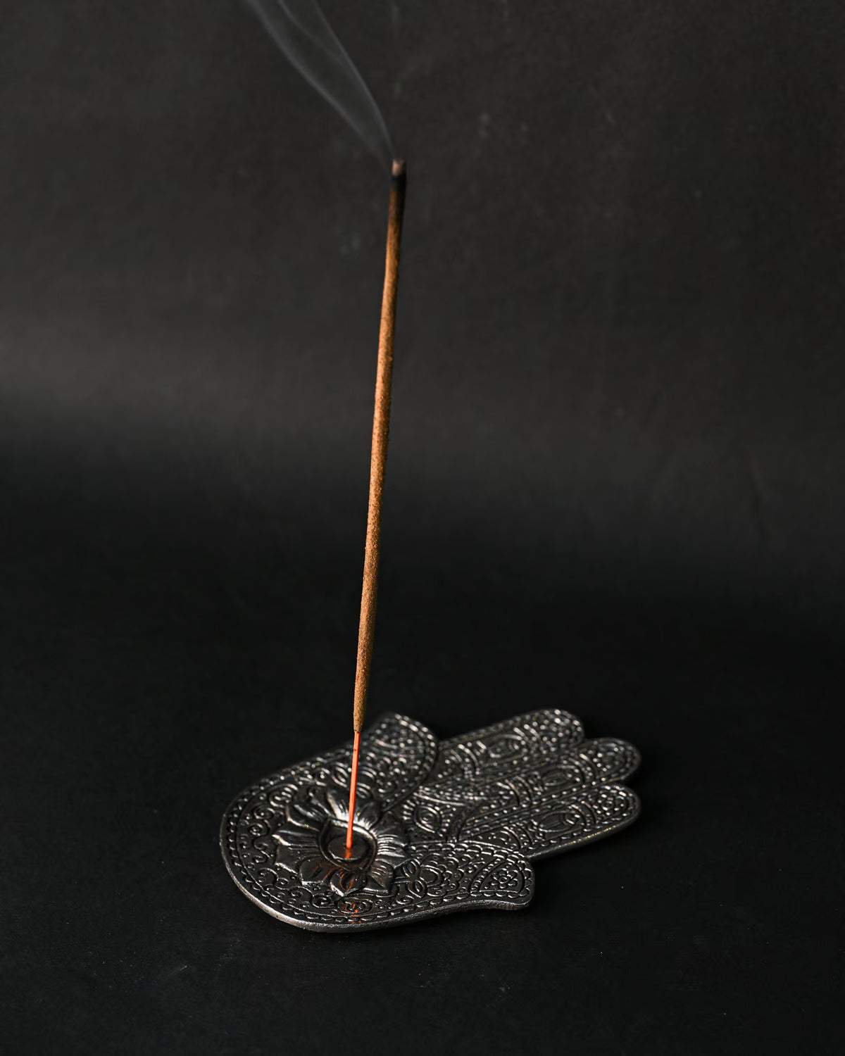 Incense stick burning upright in the hand-shaped metal incense holder, smoke rising against a dark background.
