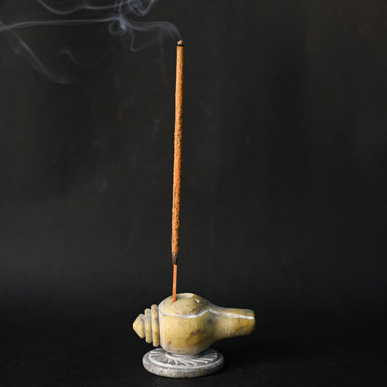 Incense stick burning upright in the conch-shaped Shankh stone holder, smoke drifting against a dark background.