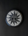 Soleil Noir incense stick holder, a round black stone disc carved with a sun face, seen from above on a dark background.