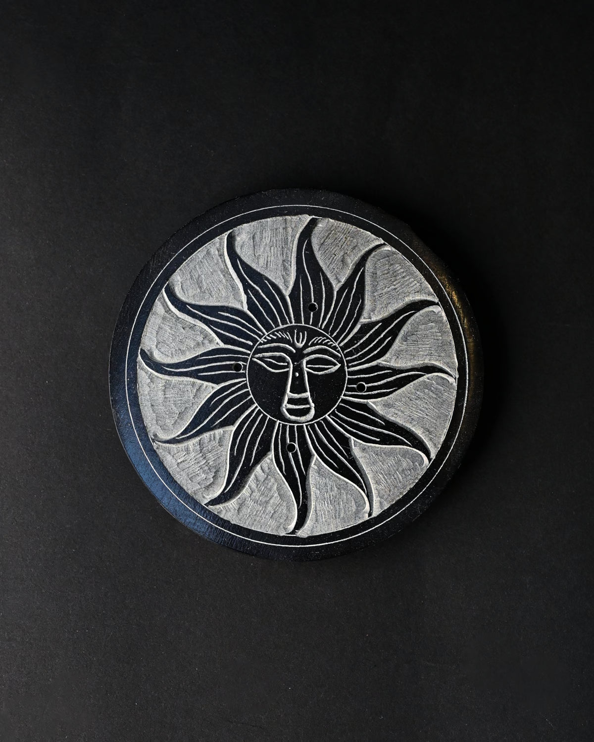 Soleil Noir incense stick holder, a round black stone disc carved with a sun face, seen from above on a dark background.