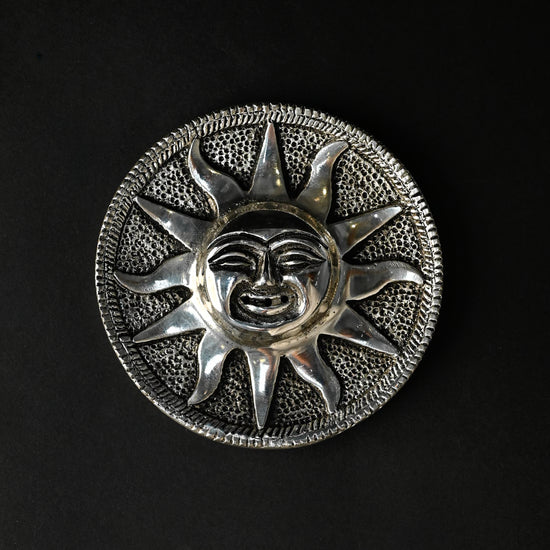 Surya incense stick holder, a round silver-toned metal plate with a sun face at its centre, on a dark background.