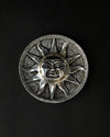 Surya incense stick holder, a round silver-toned metal plate with a sun face at its centre, on a dark background.