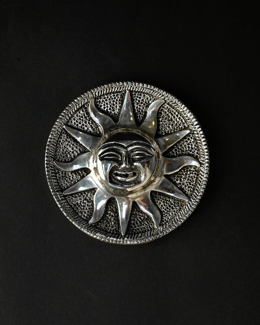 Surya incense stick holder, a round silver-toned metal plate with a sun face at its centre, on a dark background.