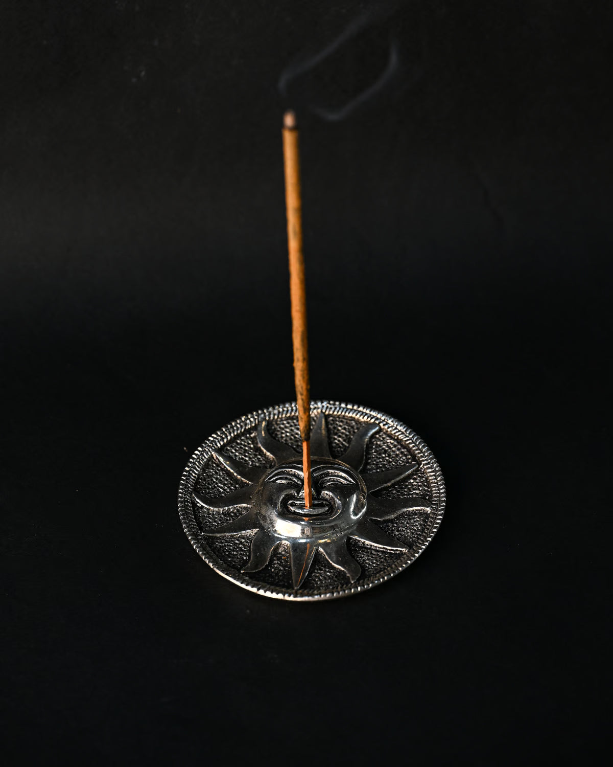 Incense stick burning upright in the Surya sun-face metal holder, smoke rising against a dark background.