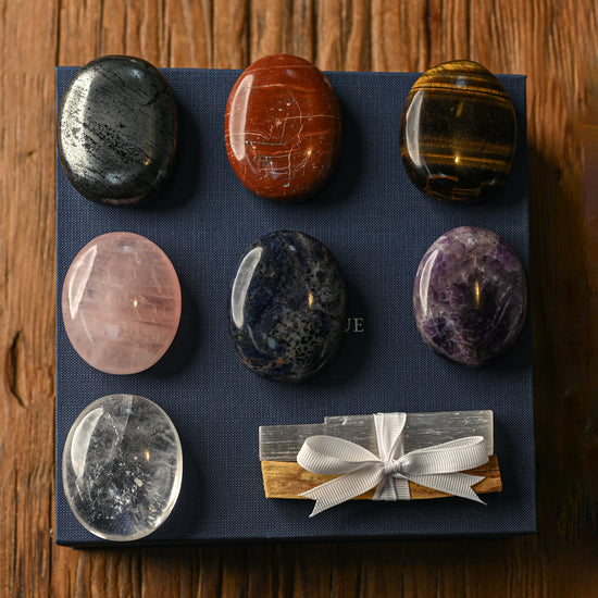 Seven polished palm stones and a ribbon-tied Palo Santo bundle arranged on a navy box, on a wooden surface.
