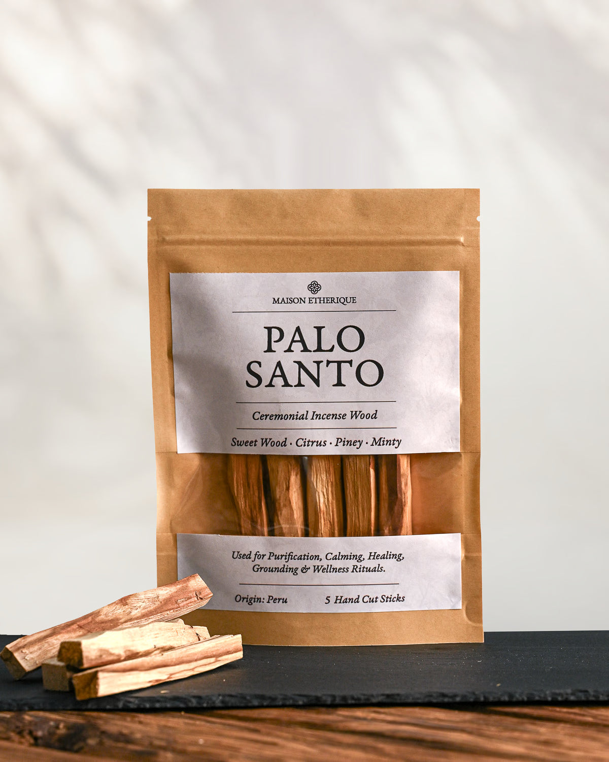 Kraft pouch of Maison Éthérique Palo Santo, five hand-cut sticks, on dark slate with loose sticks beside it.