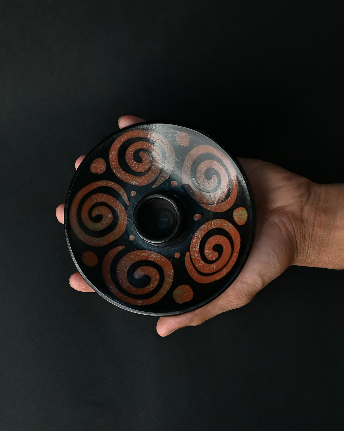 Hand holding a black ceramic Palo Santo burner painted with rust-red spirals against a dark background.