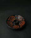 Black ceramic Palo Santo burner painted with rust-red spirals, seen at an angle on a dark background.
