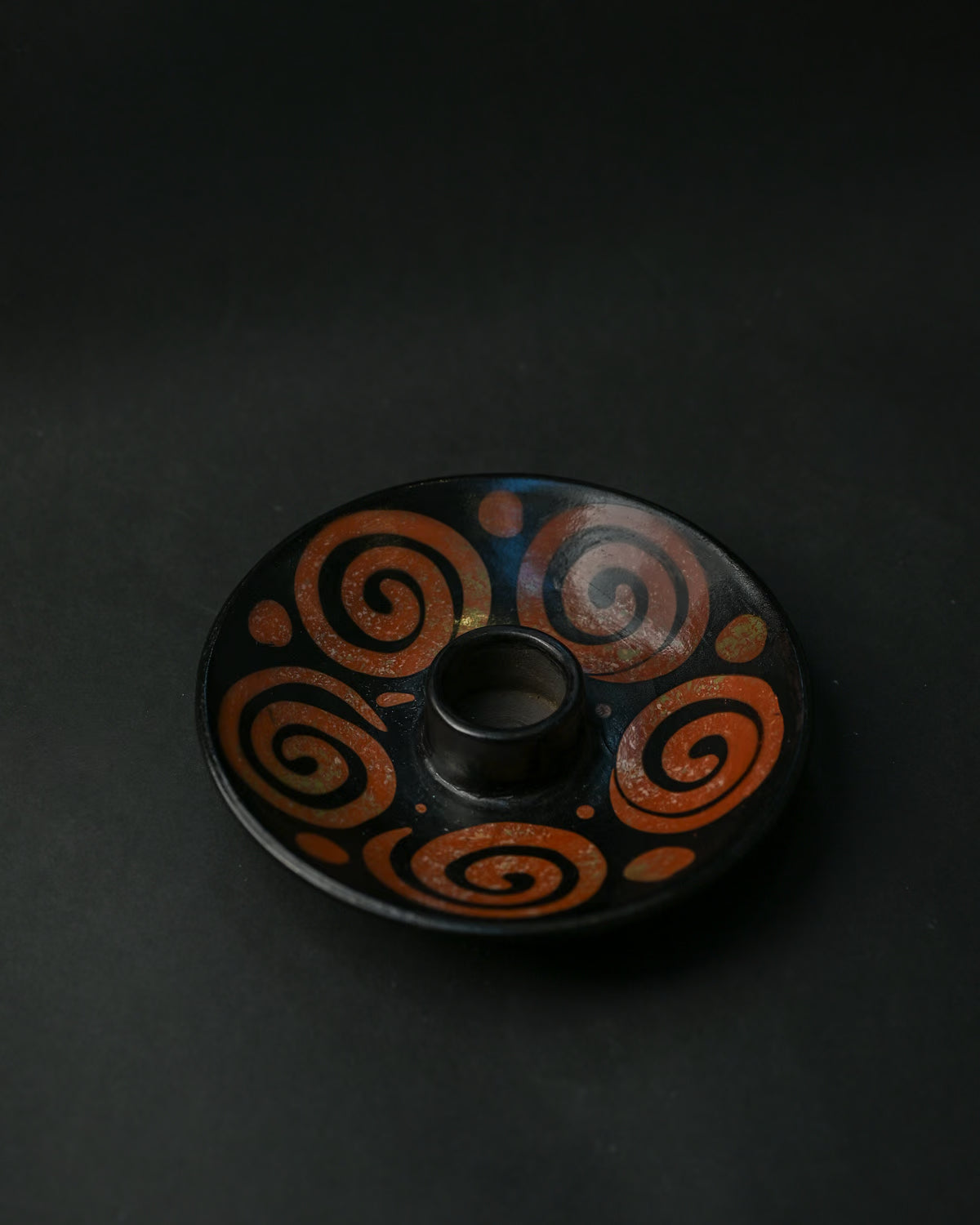 Black ceramic Palo Santo burner painted with rust-red spirals, seen at an angle on a dark background.