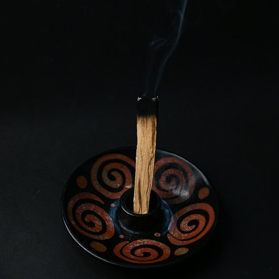 Palo Santo stick smouldering upright in a black ceramic burner painted with rust-red spirals, smoke rising.