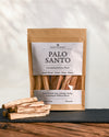Kraft pouch of Maison Éthérique Palo Santo, 100 g, standing on dark slate with loose Palo Santo sticks in front.
