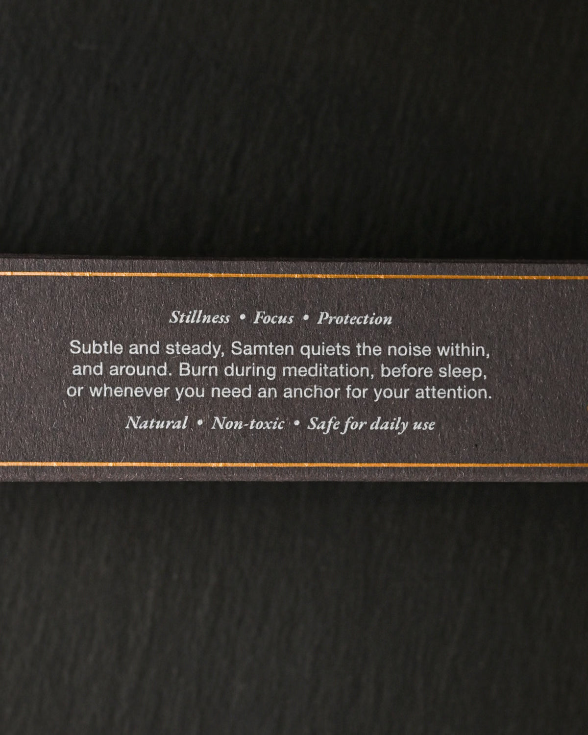 Side of the dark Samten Tibetan incense box with printed text and gold lines, on a dark background.