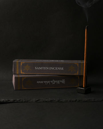 Two dark boxes of Samten Tibetan incense beside a stick burning upright in a small holder, on a dark background.