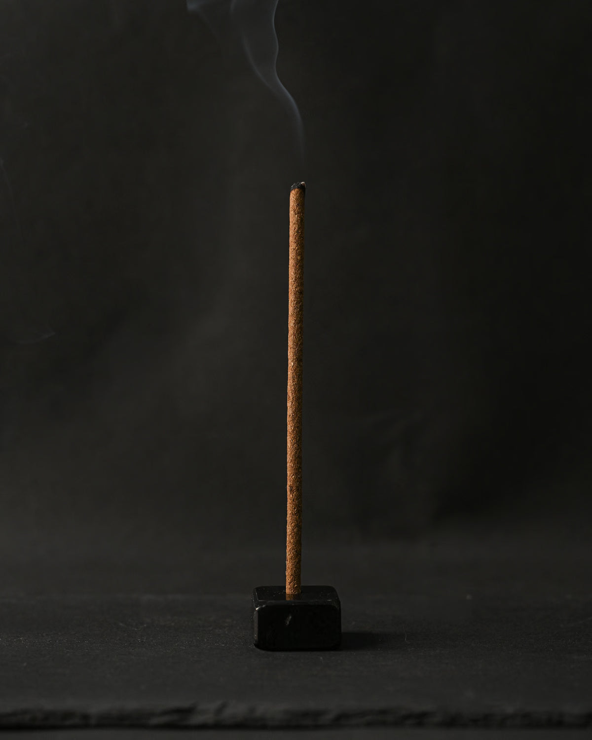 Samten Tibetan incense stick burning upright in a small dark holder, smoke rising against a dark background.