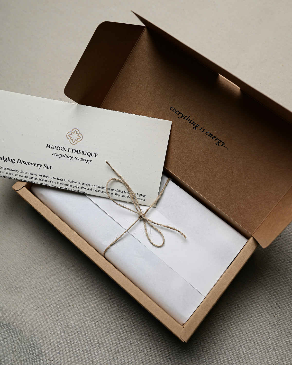 Open kraft box of the Maison Éthérique Smudging Discovery Set with a printed card tied in twine.